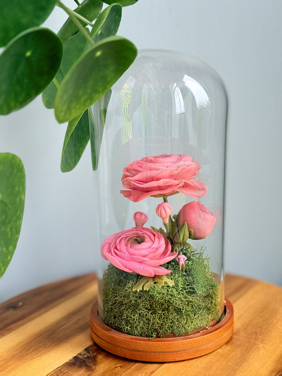 Pink ranunculus bouquet under a glass cloche atop a mossy bed and wooden base, with green leaves in the background.