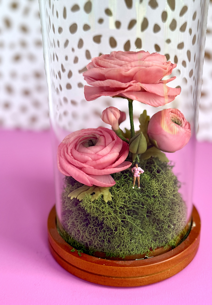 Diorama inside a glass cloche with pink ranunculus flowers, moss, and a tiny figurine on a pink base.