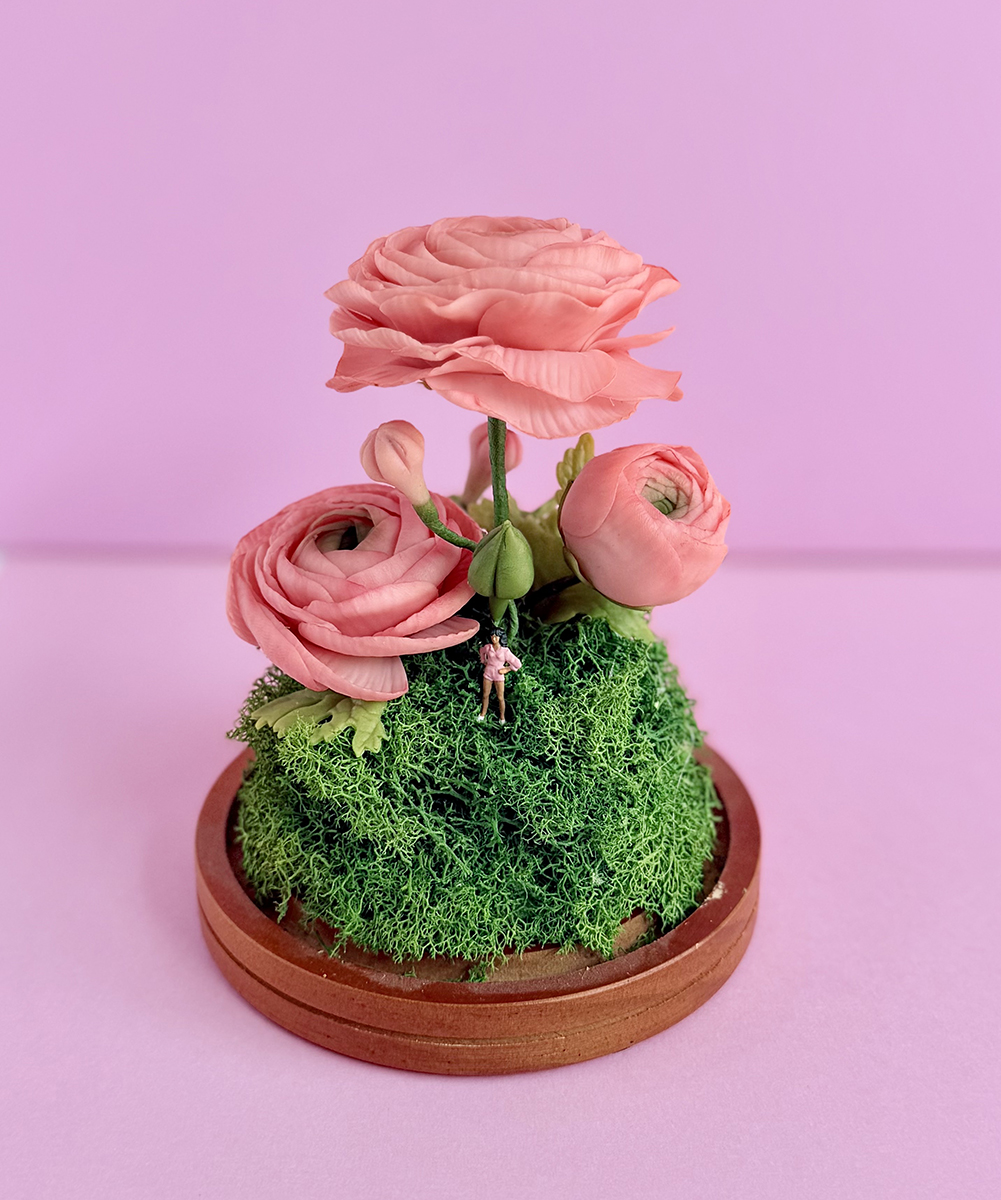 Decorative miniature bouquet of pink flowers on a mossy base with a tiny figurine, set on a wooden stand against a pink background.